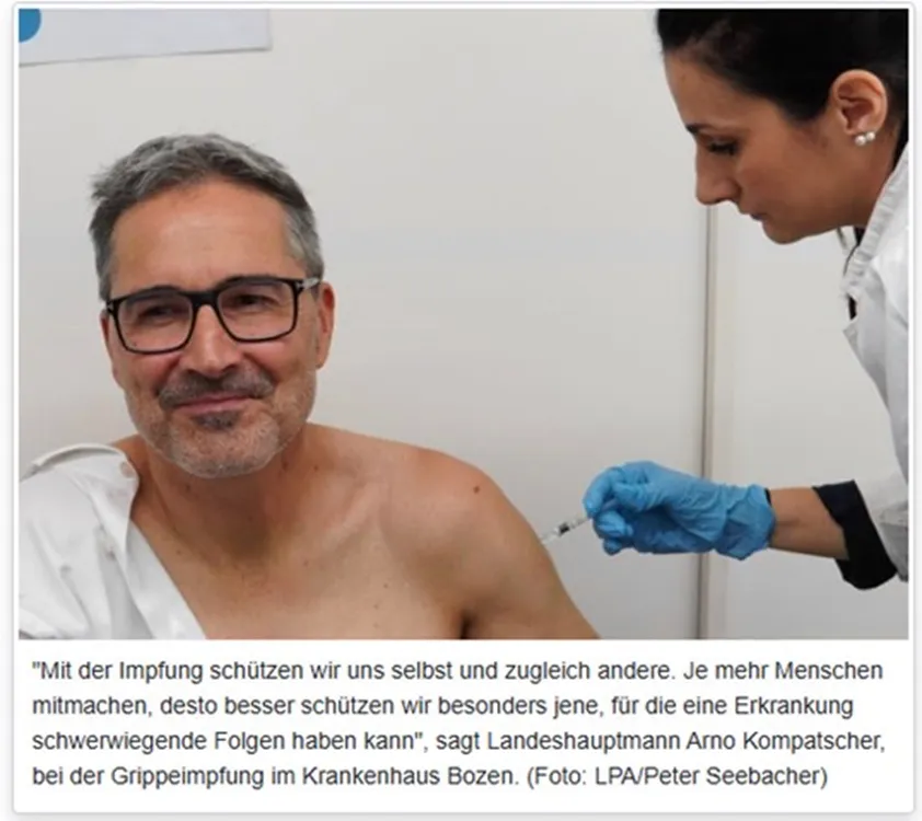 Ein Mann erhält in einem medizinischen Behandlungsraum eine Injektion in den Oberarm von einer medizinischen Fachkraft mit blauen Handschuhen.