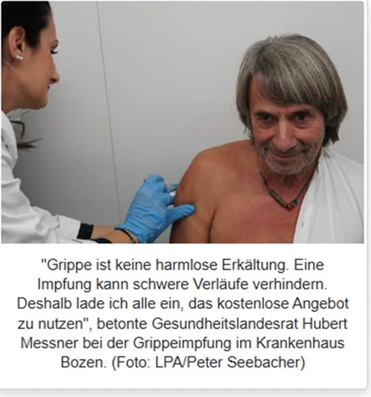 Ein Mann erhält in einem medizinischen Behandlungsraum eine Injektion in den Oberarm von einer medizinischen Fachkraft mit blauen Handschuhen.