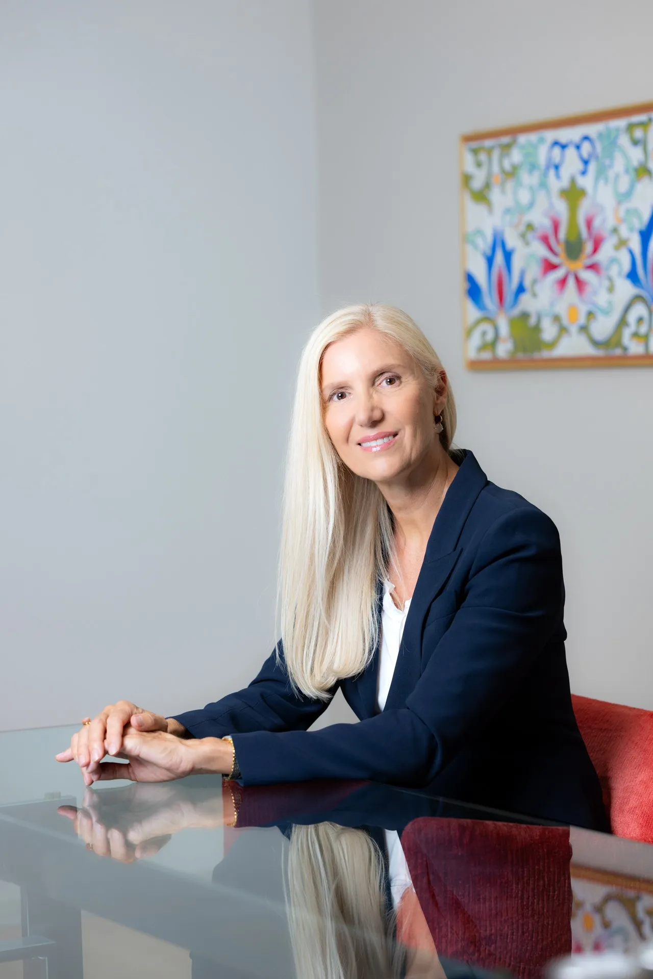 Eine lächelnde Frau mit langen blonden Haaren im dunkelblauen Blazer sitzt an einem Tisch.